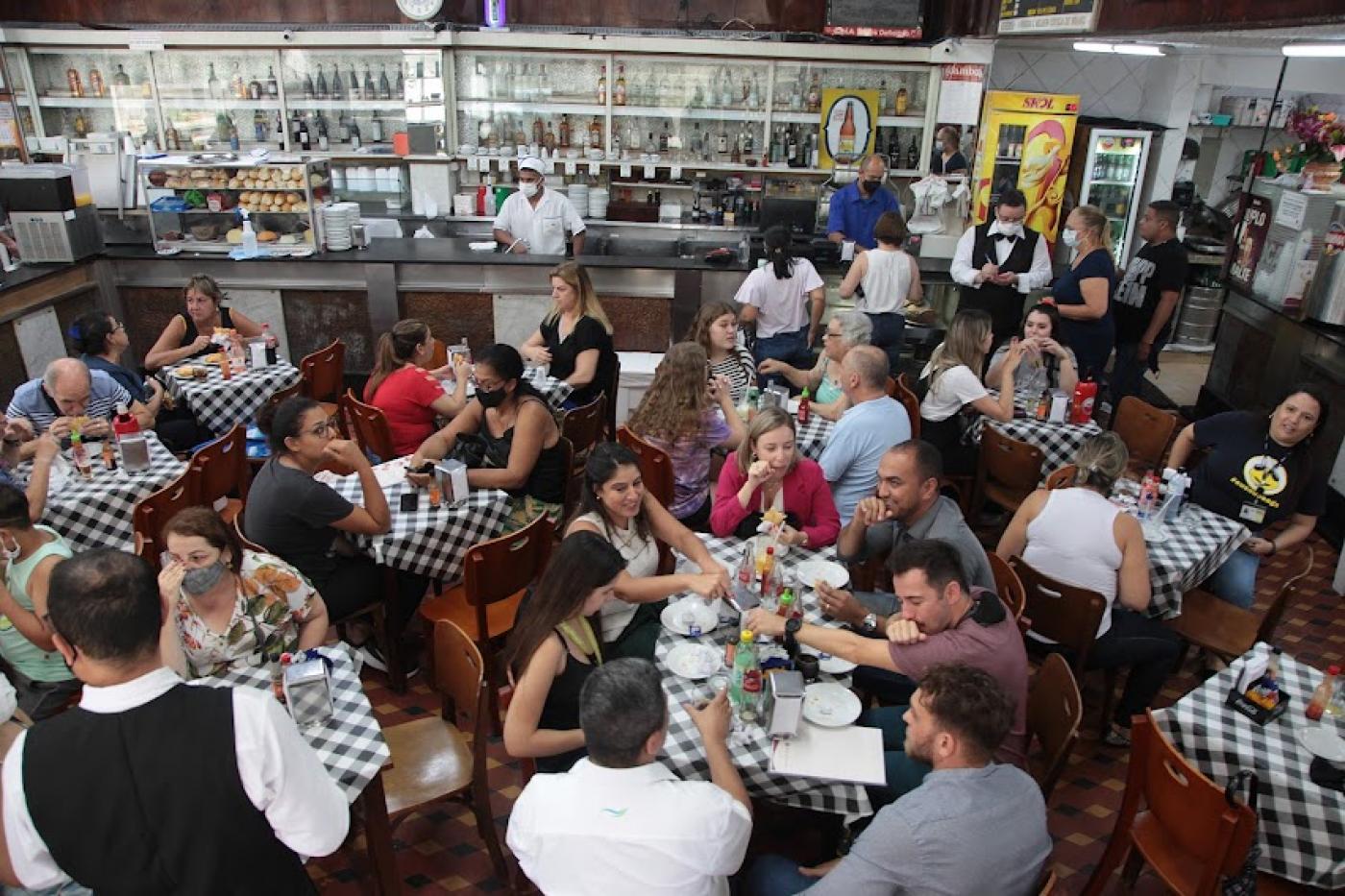 Restaurante lotado de clientes sentados em torno de mesas com toalhas estampadas. #paratodosverem
