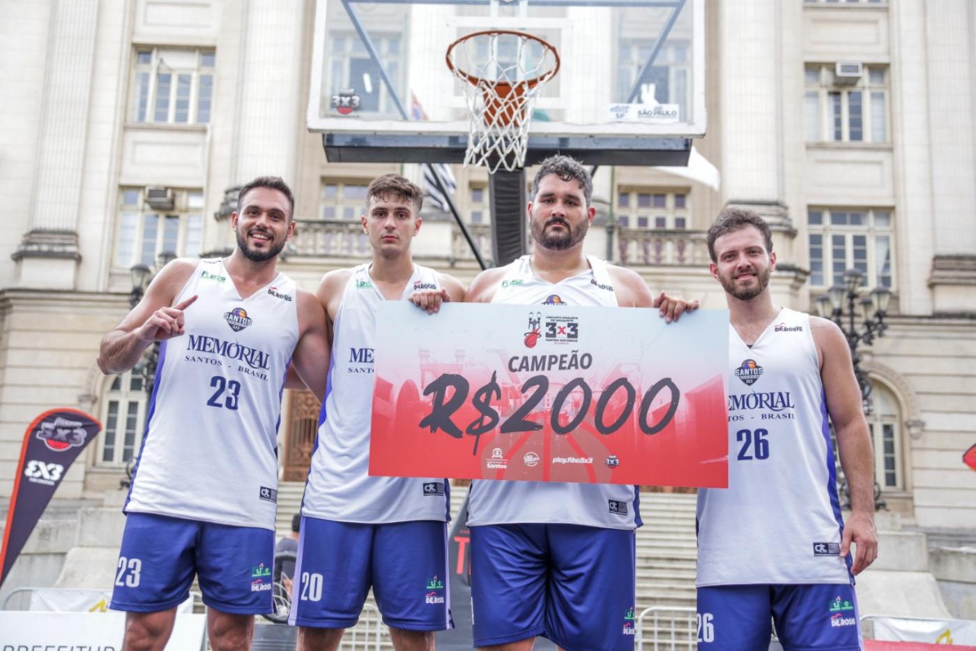 Atletas mostram cheque de R$ 2 mil da premiação em frente à cesta de basquete, com Prefeitura ao fundo. #pracegover