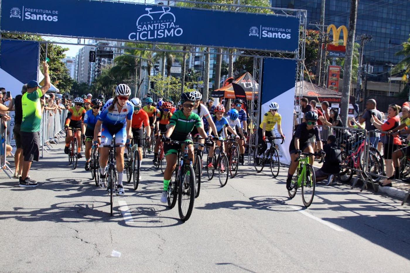 ciclistas estão fazendo largada de prova. Pórtico ao fundo tem escrito Campeonato Santista de Ciclismo. #paratodosverem