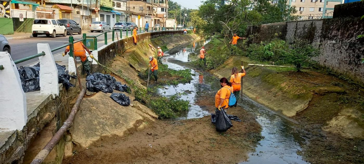 homens e mulheres uniformizados estão dentro de canal com pás e sacos plásticos. Às margens se vê terra e vegetação. #paratodosoverem