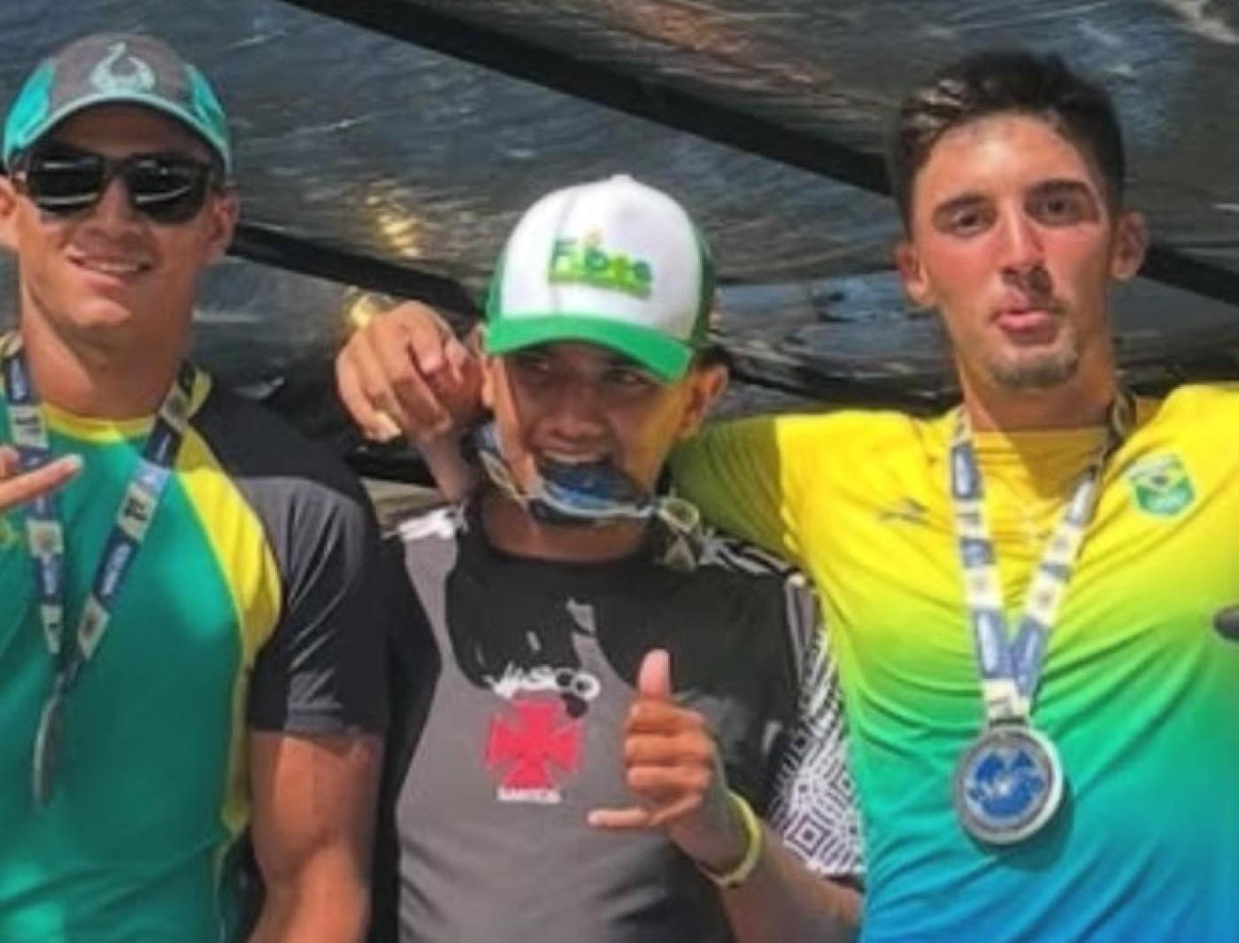 trés atletas posam para foto abraçados. O do meio segura uma medalha
