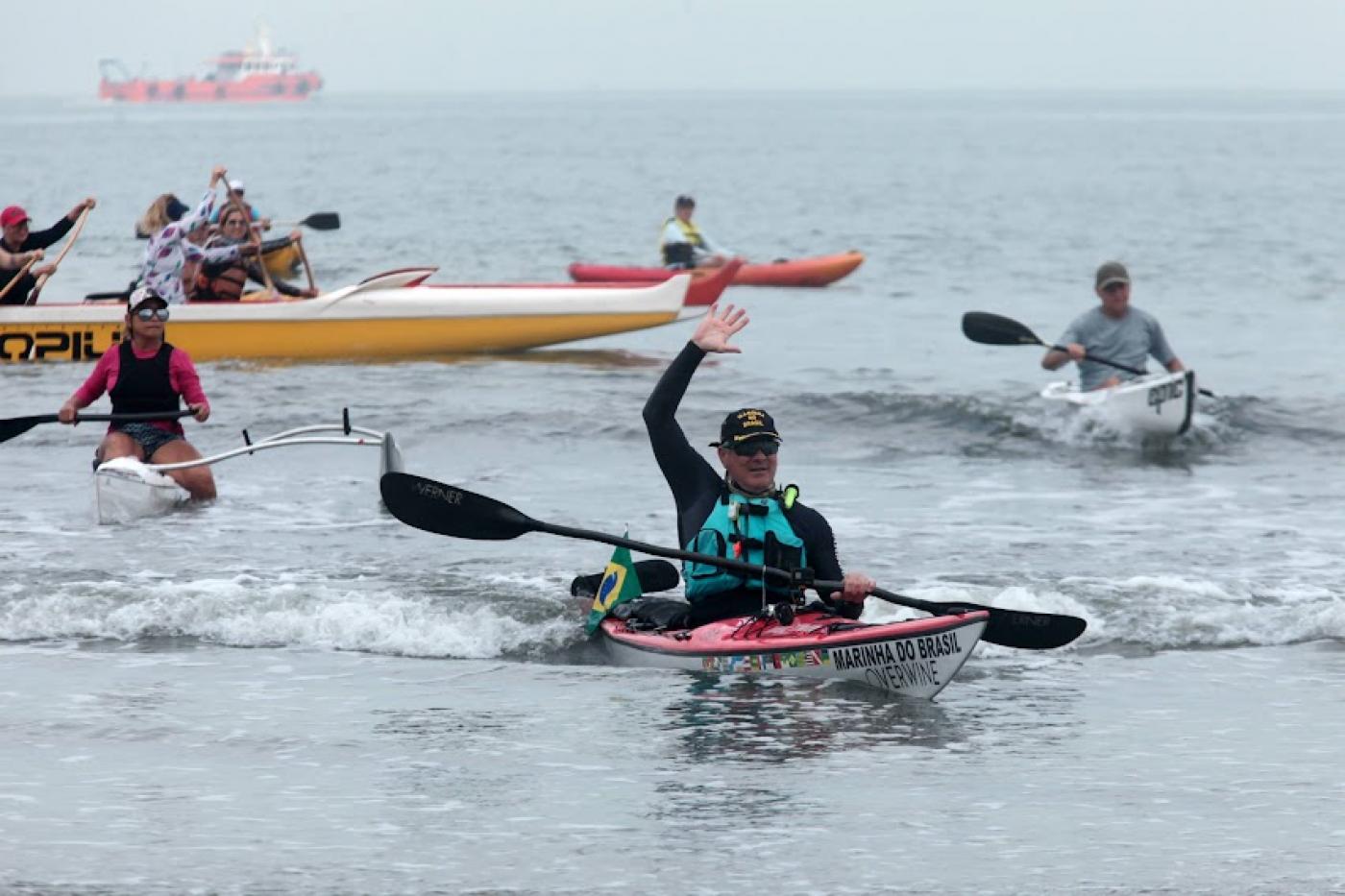 canoísta está dentro do caiaque na beira do mar. Com uma mão ele segura o remo e outra está erguida para o alto. #paratodosverem