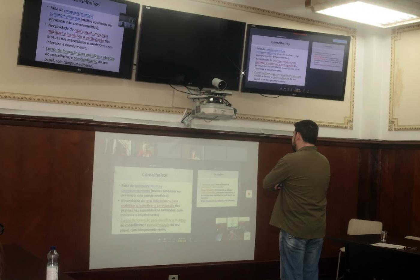 homem está em pé em frente a telão afixado em parede. ele está dando um curso on-line. #paratodosverem