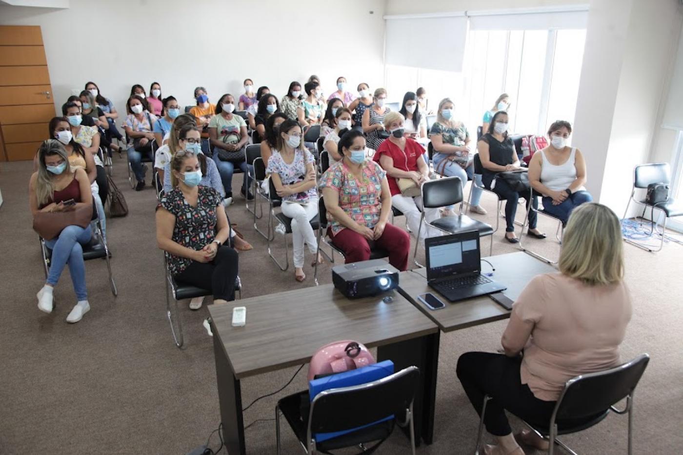 profissionais de saúde estão sentados em sala, em cadeiras distantes umas das outras. Ele olham para mulher que está sentada diante uma mesa com um notebook. #paratodosverem