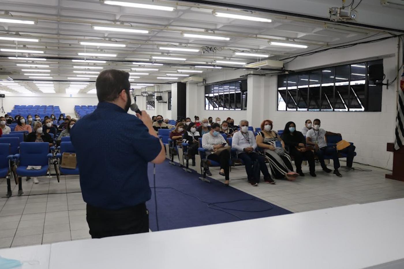 homem está falando ao microfone e à frente de uma mesa. Ele está de costas para a foto. Várias pessoas estão sentadas no auditório. #paratodosverem