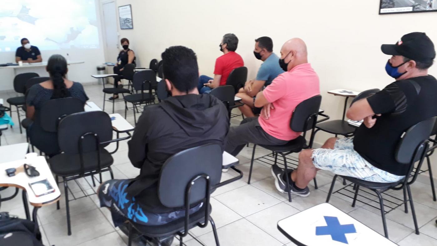 Agentes sentados em cadeiras observam instrutor com telão ao fundo. #pratodosverem