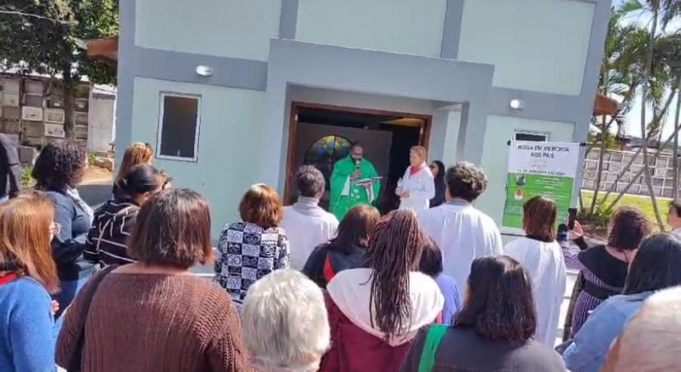 pessoas em pé diante de capela assistem padre falando ao fundo. #paratodosverem 