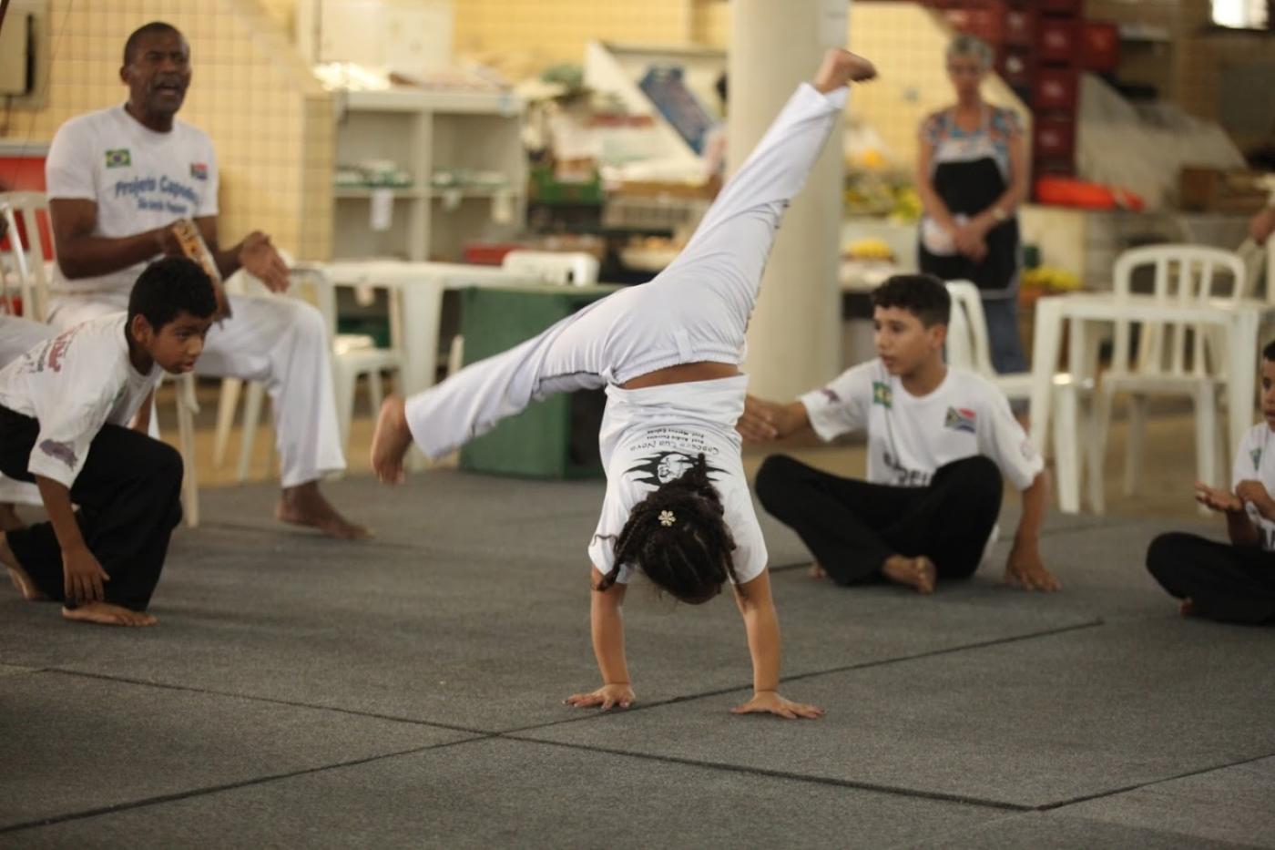 menina está dando estrela em aula de capoeira. Suas pernas estão abertas no ar. As duas mãos no chão. Ao fundo, mais crianças agachadas. 