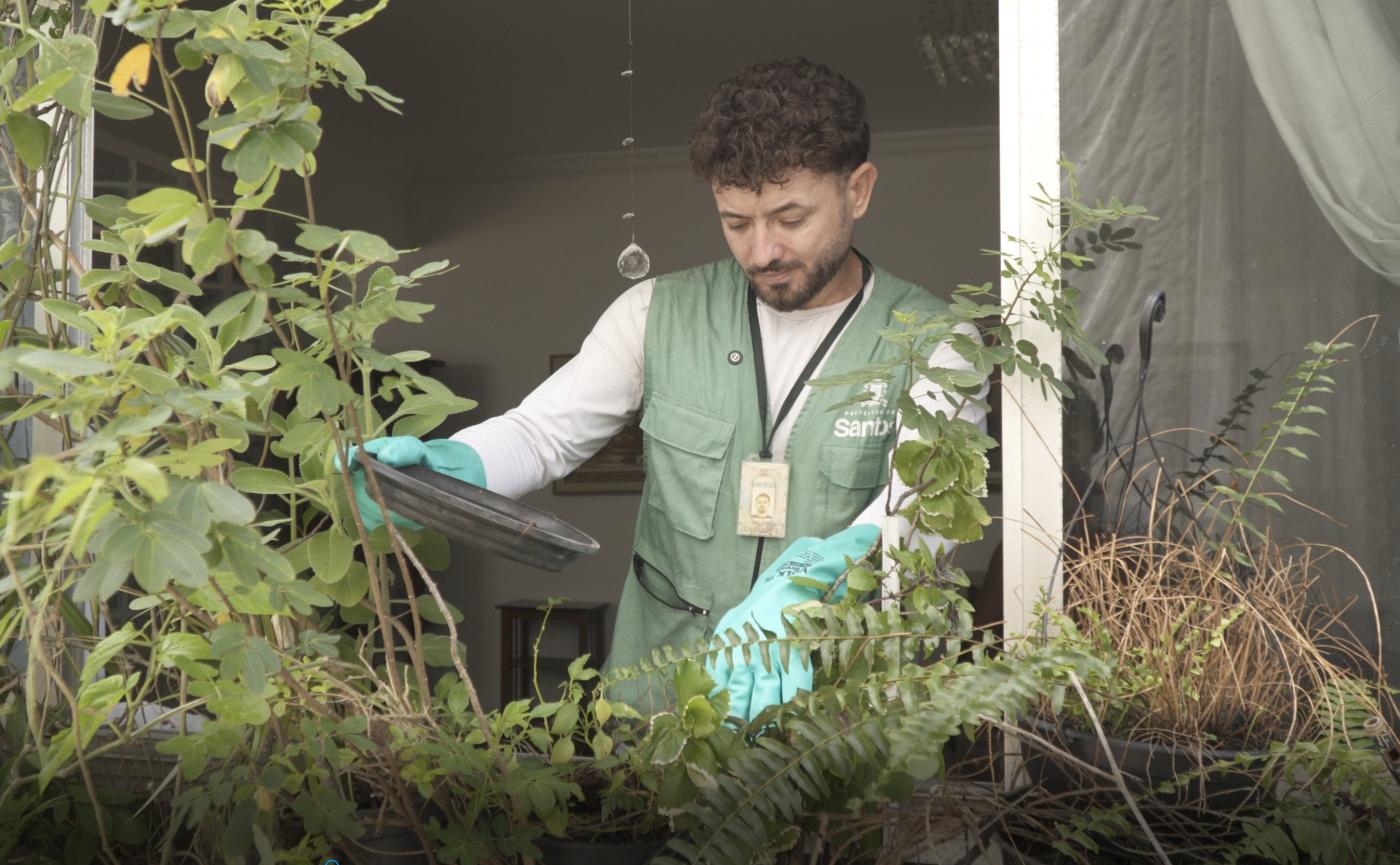 agente de combate à dengue está atrás de janela aberta mexendo em plantas voltadas para fora. Ele usa luvas. #paratodosverem 