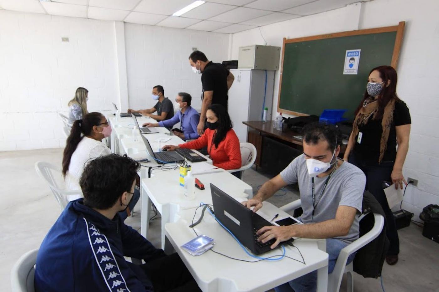 pessoas estão sentadas diante de mesas e são atendidas por outras pessoas que estão com laptops abertos. #paratodosverem