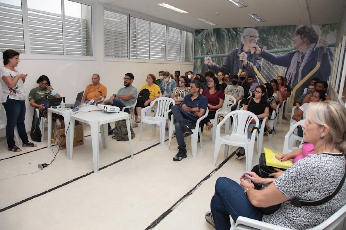 sala com pessoas sentadas e uma mulher em pé falando a todos. #paratodosverem