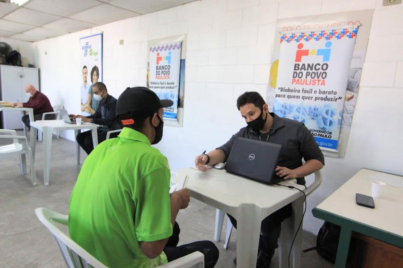 homem está sentado com laptop aberto à sua frente. Ele atende outro homem que está de costas para a foto. Ao fundo, um banner do banco do povo. #paratodosverem