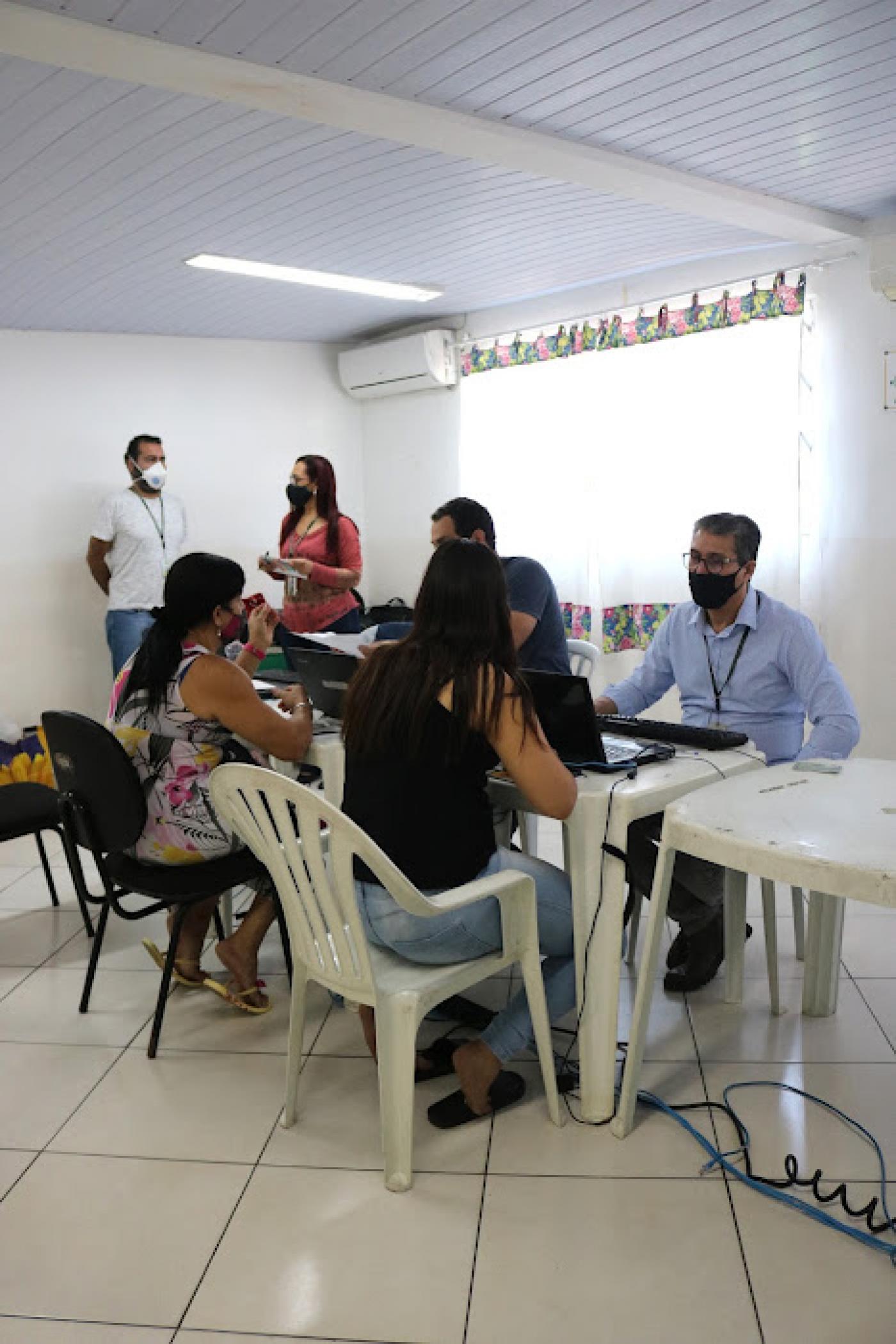 Mulheres, de costas, são atendidas por profissionais sentados de frente para computadores. #pratodosverem
