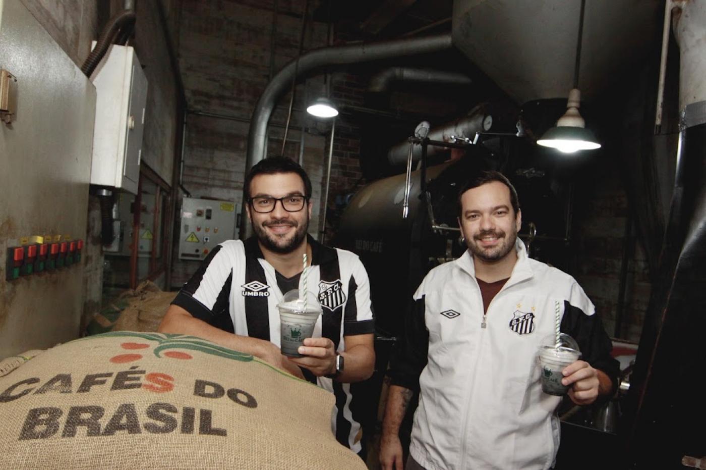 dois jovens com camisa do santos segurando café com saco de fé a frente e maquinário ao fundo 