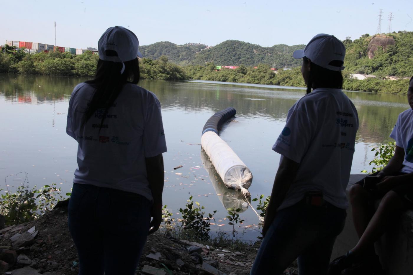 duas pessoas olham para trecho de mar com barreira flutuante. #paratodosverem 