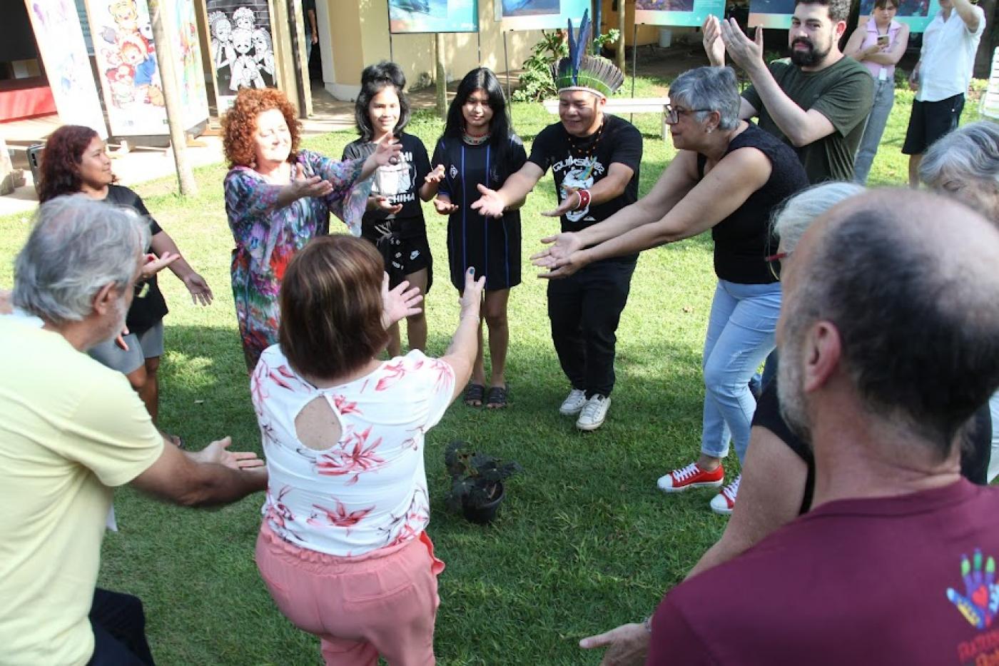 diversas pessoas formam uma roda ao ar livre e movimentam os braços para cima. #paratodosverem 