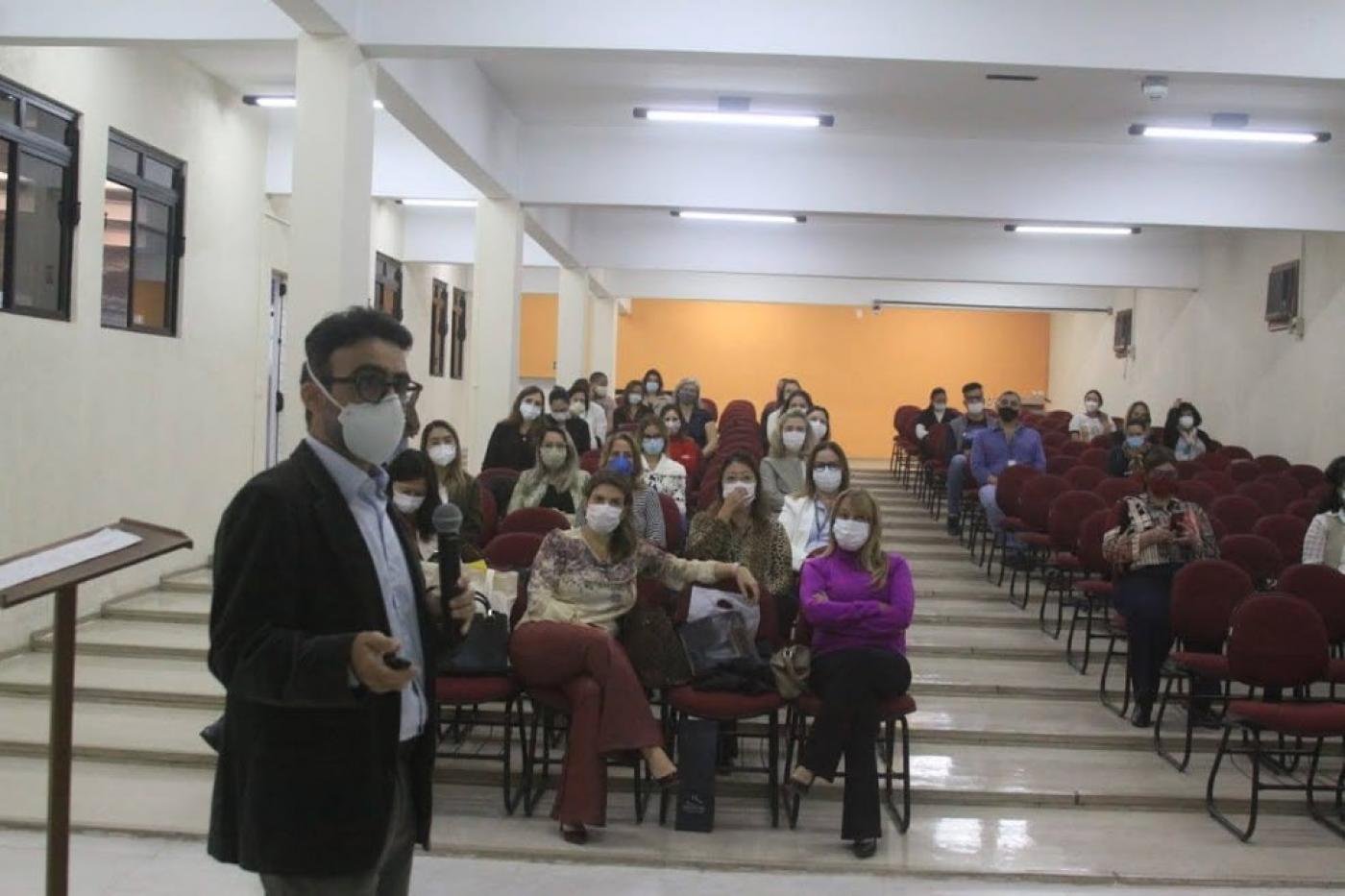 Homem em primeiro plano dando palestra e atrás mulheres sentadas em cadeiras. #paratodosverem