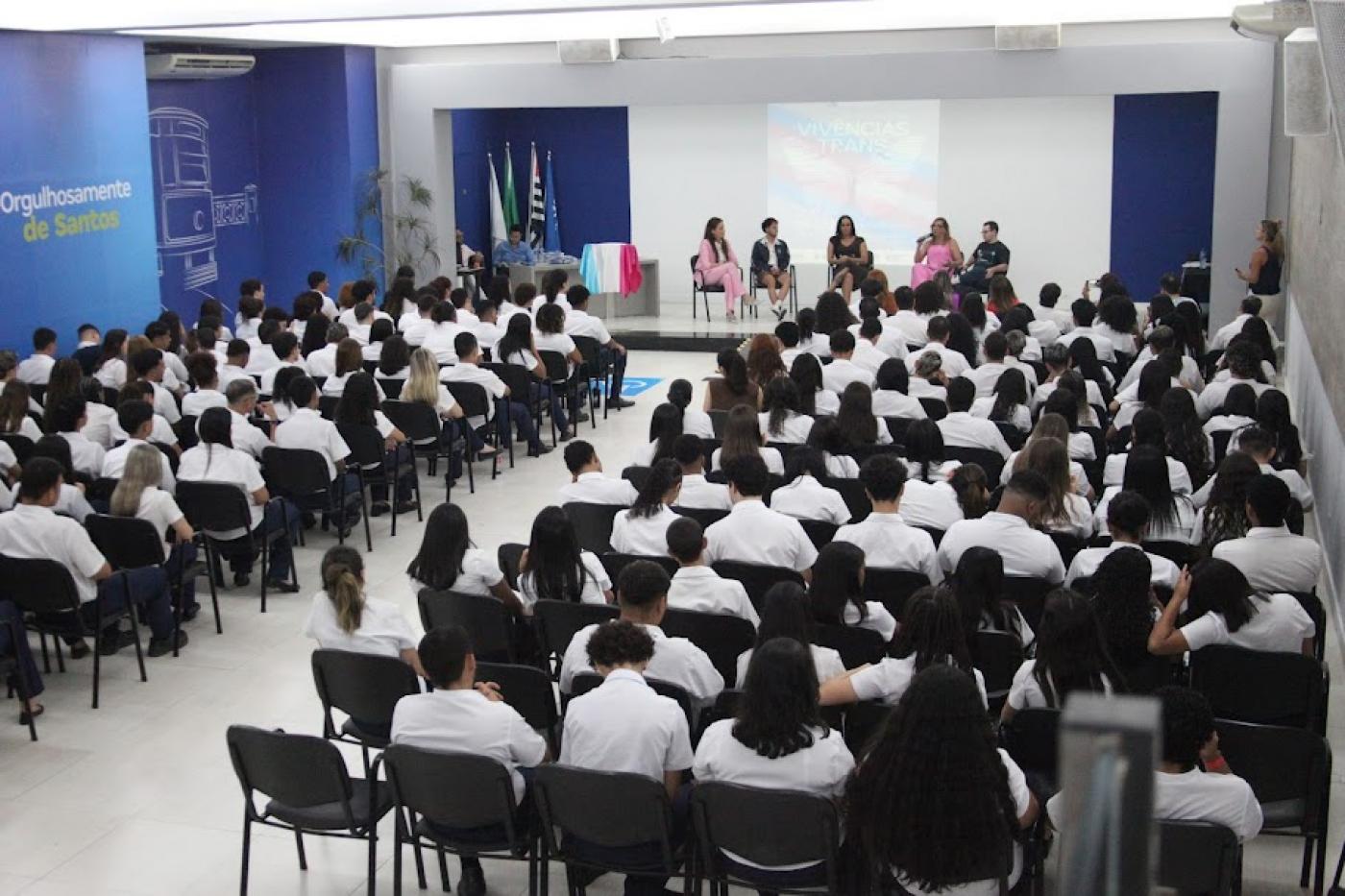 várias pessoas sentadas em auditório assistindo palestra com cinco pessoas no palco ao fundo. #paratodosverem
