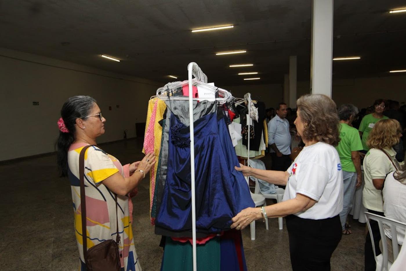 duas mulheres estão mexendo em roupas penduradas em arara num saguão. #paratodosverem 