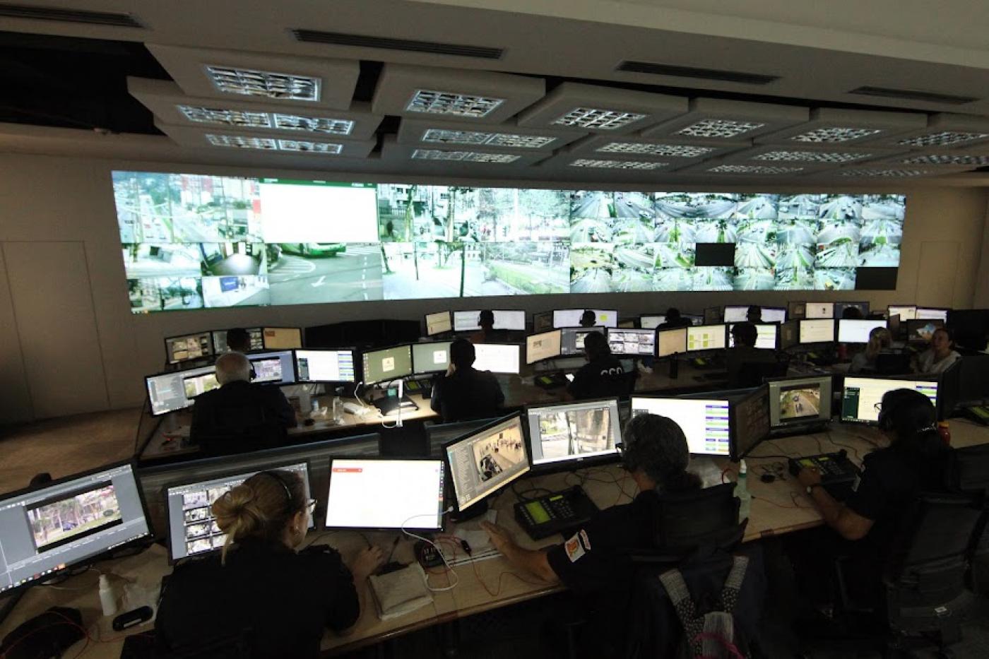 centro de controle operacional com vários telões com imagens de pontos da cidade e pessoas operando computadores em primeiro plano. #paratodosverem 