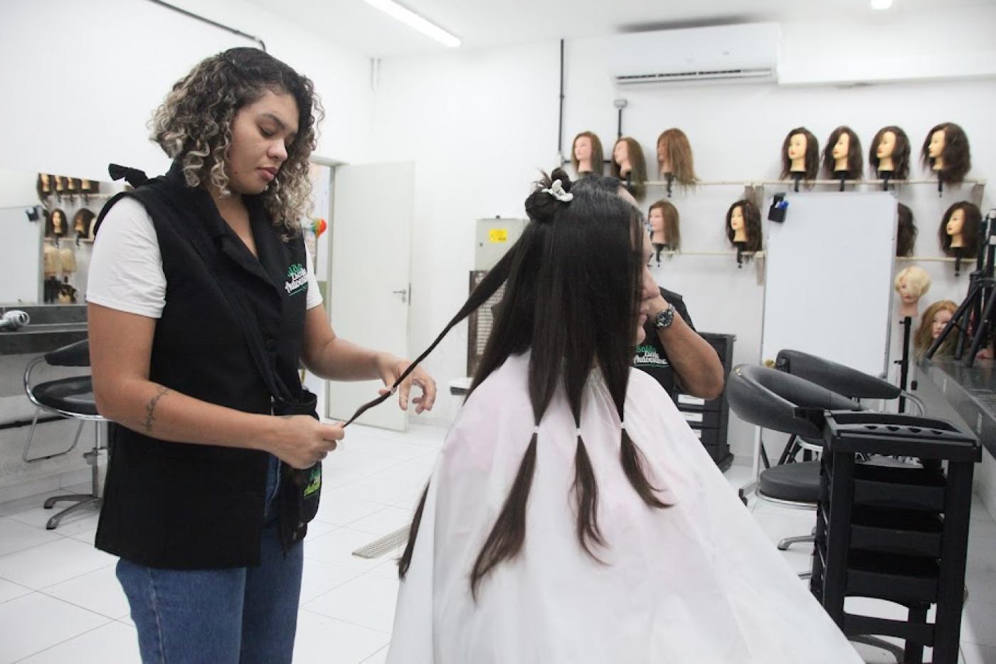 mulher está em pé cortando mechas de cabelo de uma mulher que está sentada. #paratodosverem