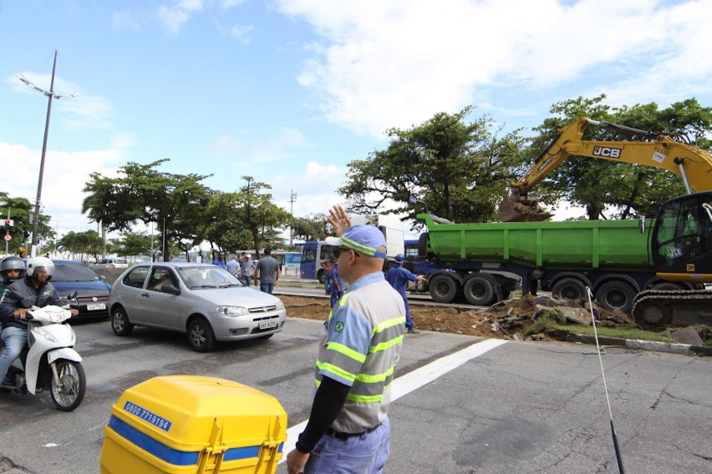 agente de trânsito está sinalizando para veículos. Ao fundo, no canteiro central há um caminhão e uma escavadeira. #paratodosverem