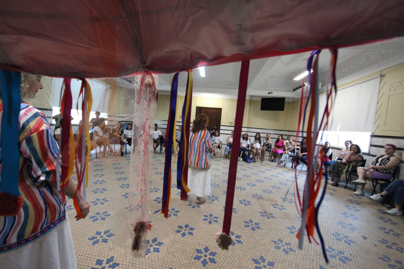Tenda com fios coloridos em primeiro plano e mulheres sentadas em roda ao fundo. #paratodosverem 
