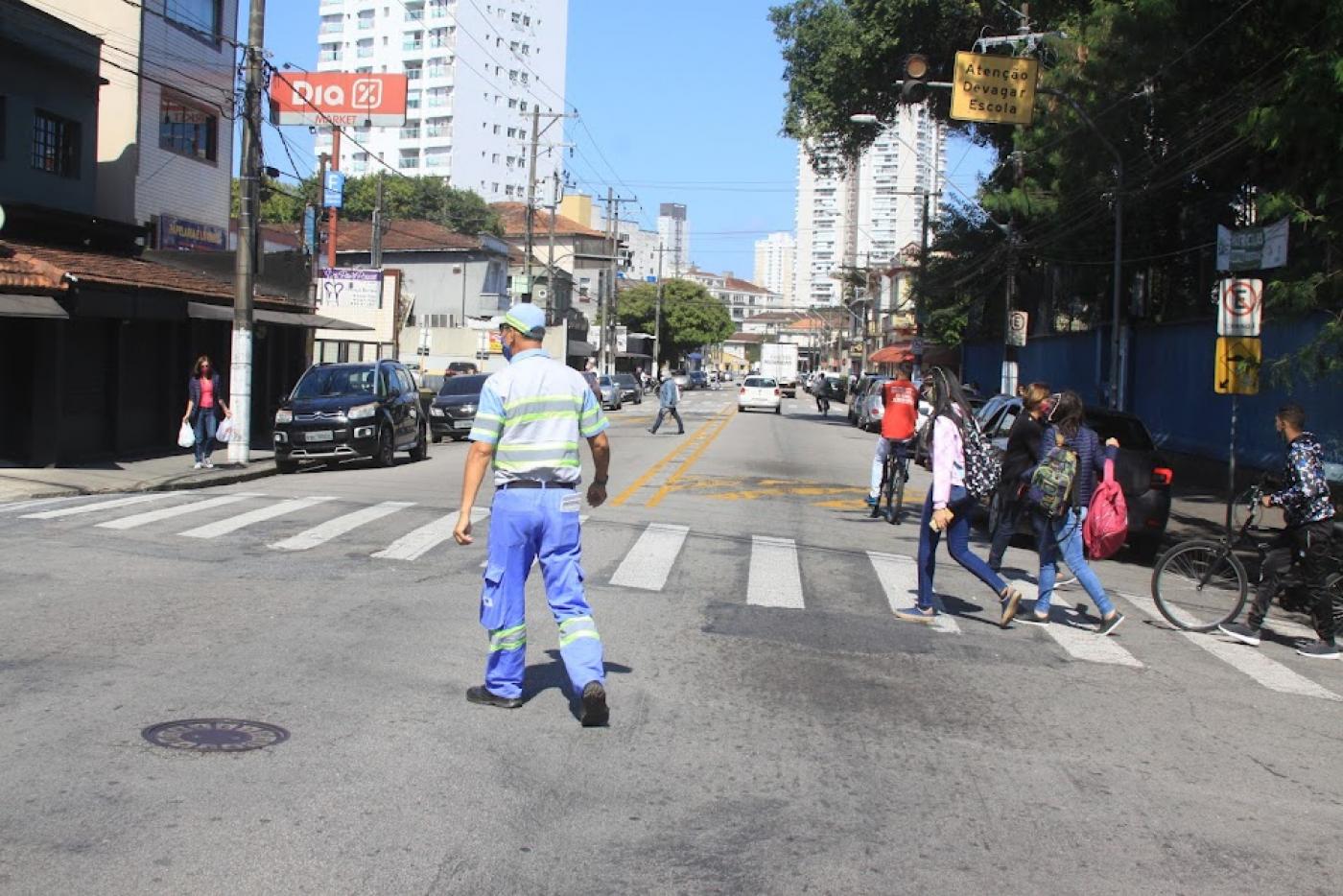 agente está no meio de uma rua, de costas para a foto e de frente para faixa de pedestres. Algumas pessoas cruzam a rua. #paratodosverem