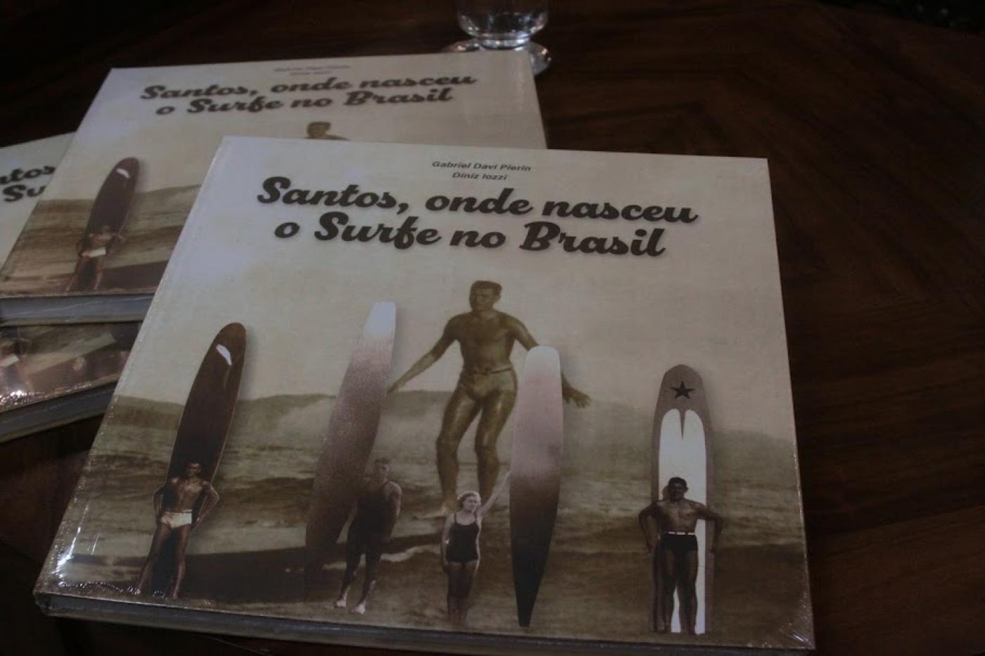 livros sofre a mesa. a foto é de alguns surfistas na praia com pranchões antigos. no alto se lê Santos, onde nasceu o surfe no Brasil. #paratodosverem 