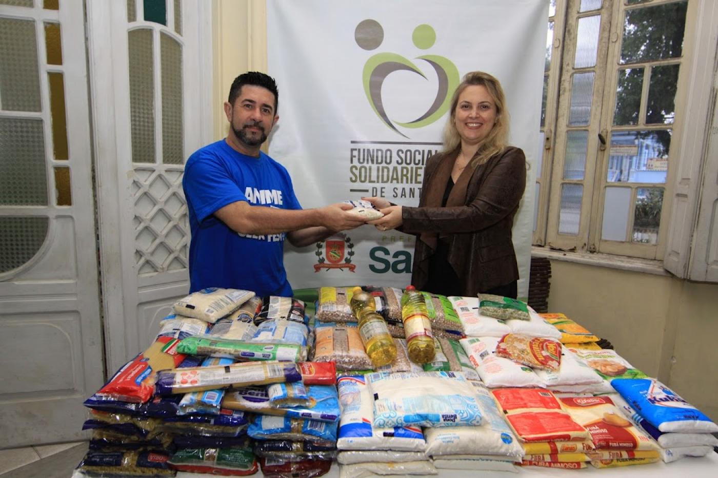 organizador do evento entrega alimento para representante do fundo com vários alimentos em mesa #paratodosverem