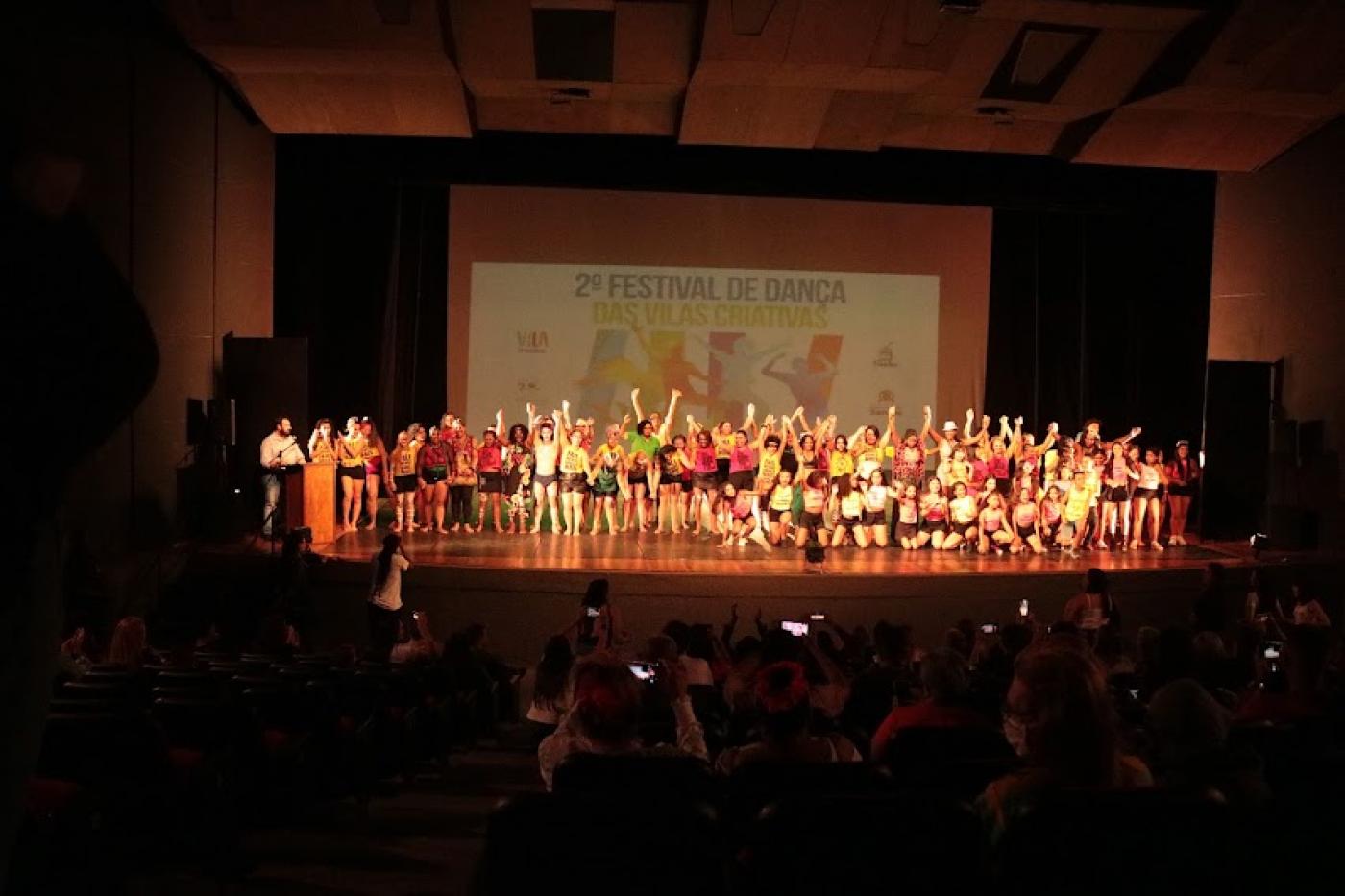 público de costas para a foto fotografando e aplaudindo. ao fundo, no palco, todos os atores da apresentação reunidos. #sparatodosverem