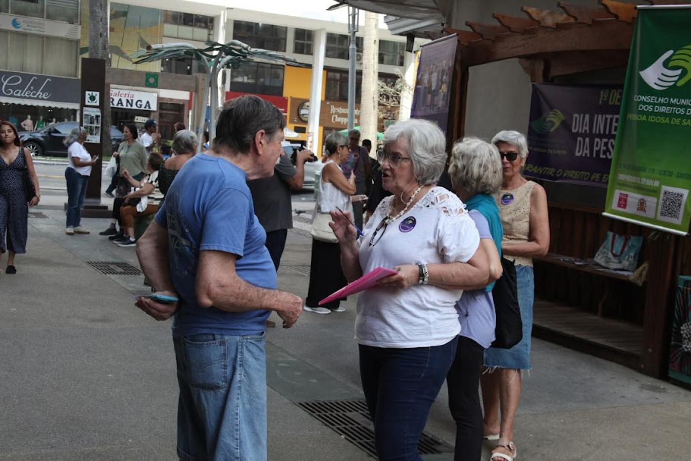 mulher segurando pracheta está conversando com homem. Ao fundo, há banner pendurado com informações para o segmento da terceira idade. #paratodosverem 