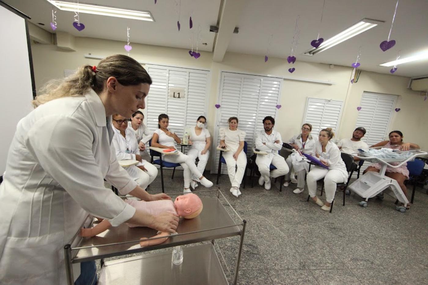 mulher massageia boneco diante de mulheres que estão sentadas assistindo. #paratodosverem 