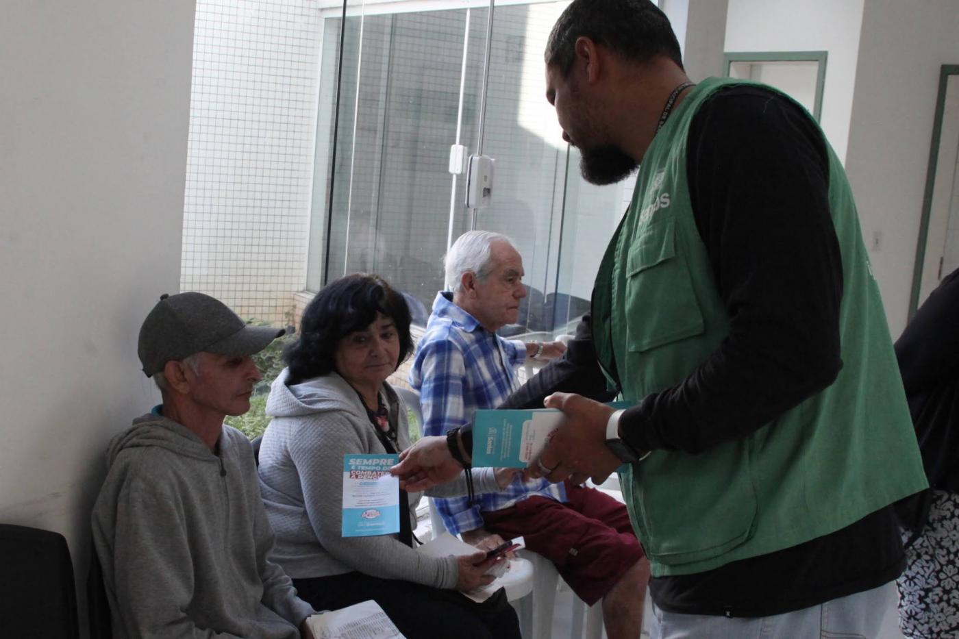 homem entrega folheto a pessoas que estão sentadas. #paratodosverem
