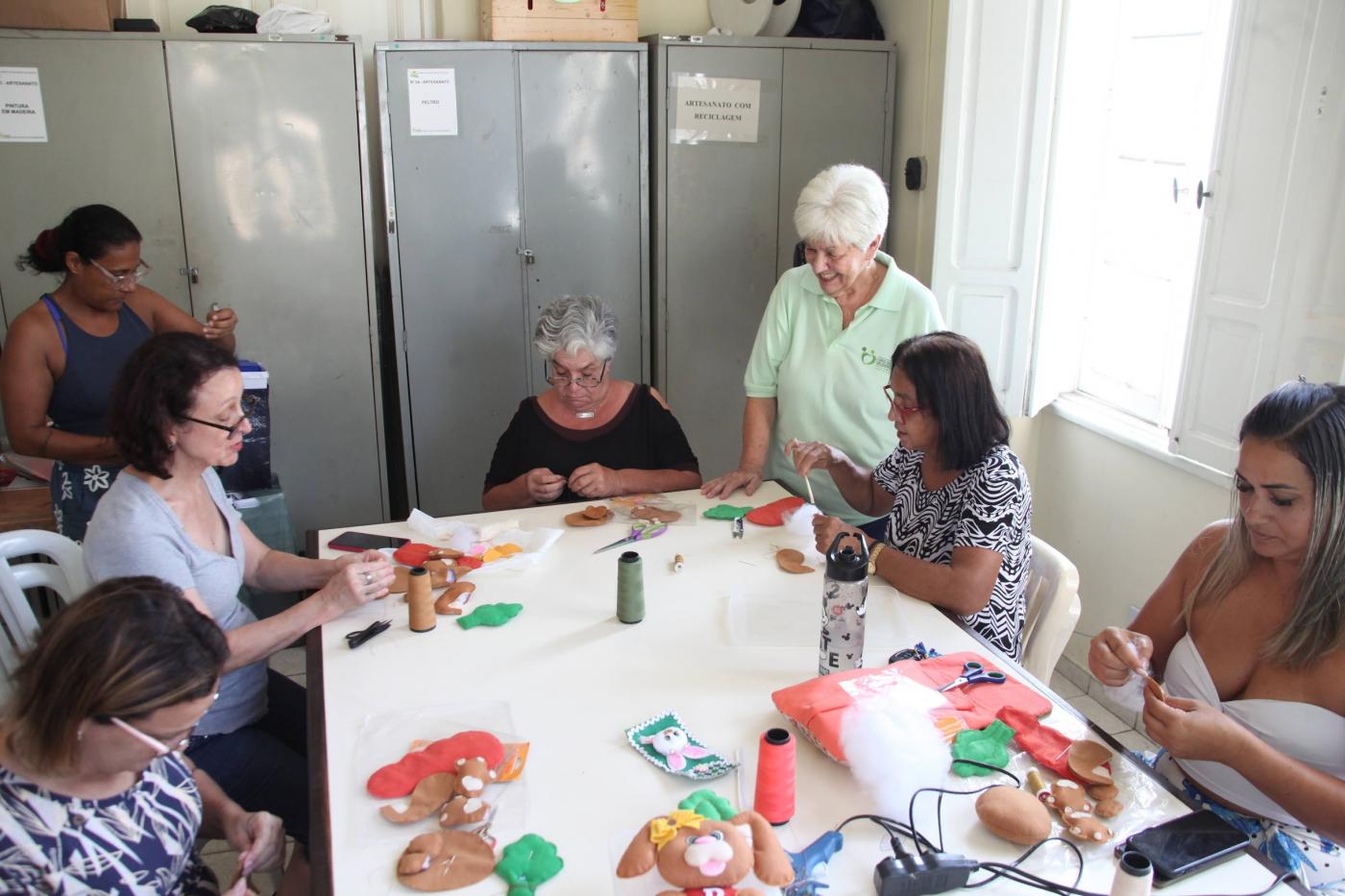 mulheres sentadas em torno de mesa fazem artesanato. #paratodosverem 