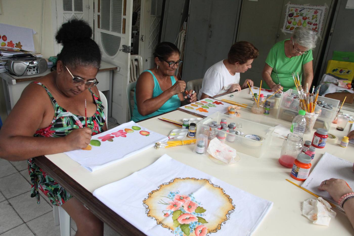 mulheres pintam panos de prato sobre uma mesa. #paratodosverem