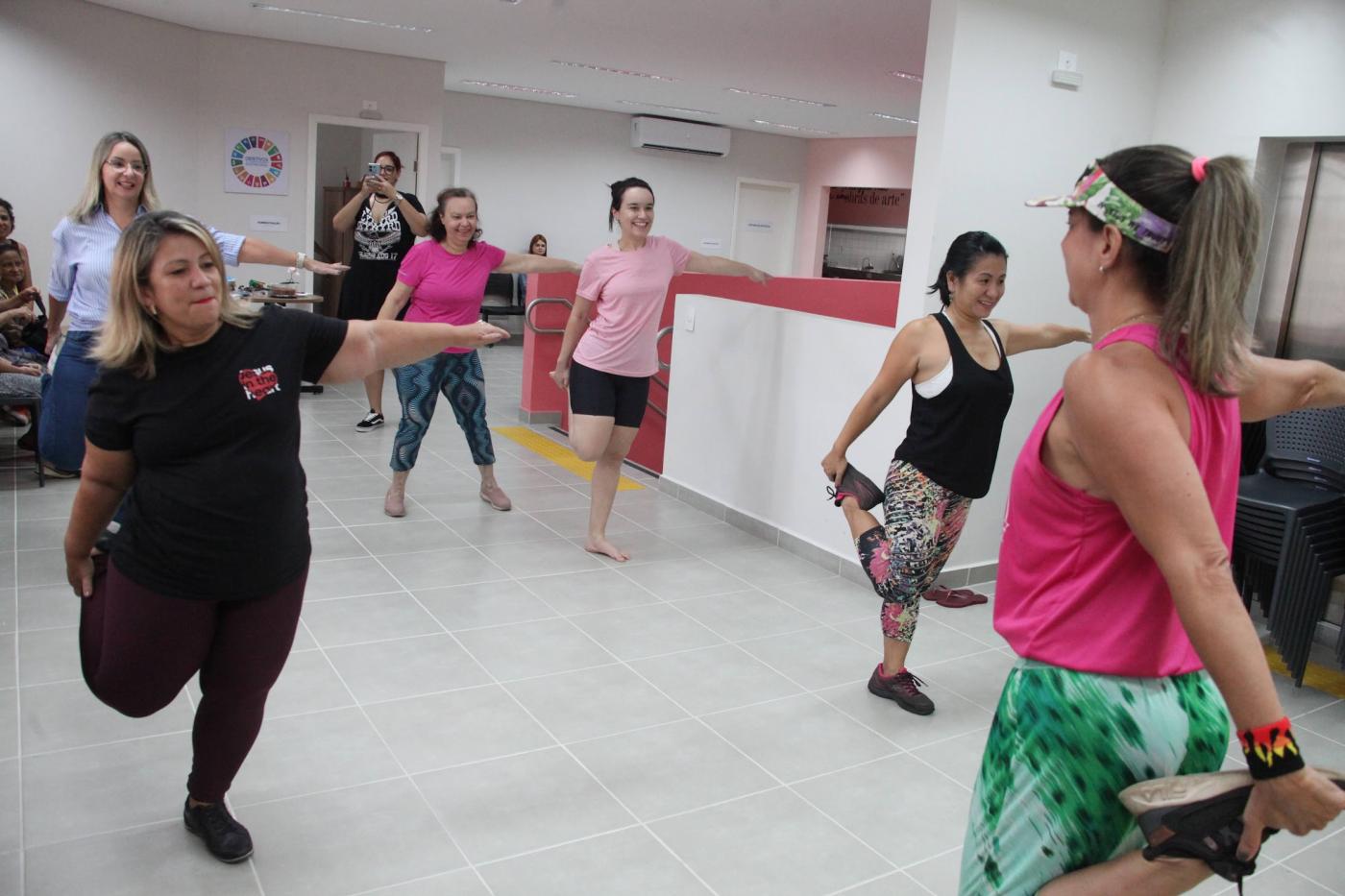mulheres fazem alongamento com as pernas em salão. #paratodosverem 