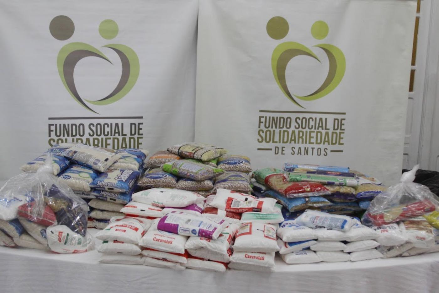 pacotes de alimentos sobre uma mesa como banner do fundo social ao fundo. #paratodosverem