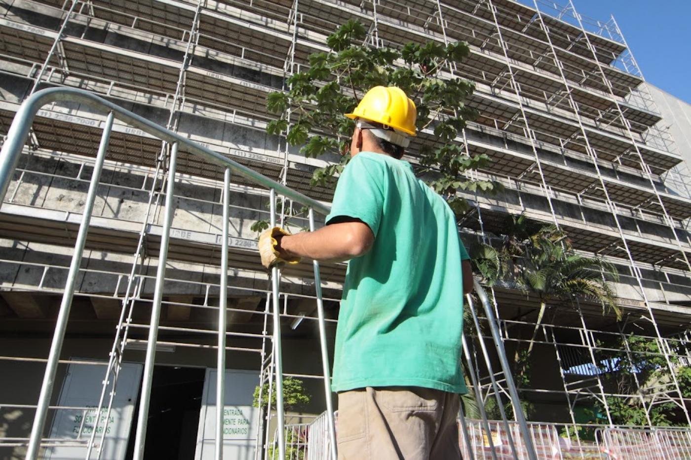 homem segura estrutura de ferro diante de fachada lateral de prédio. Ele usa capacete e luva. Está de costas para a foto. #paratodosverem 