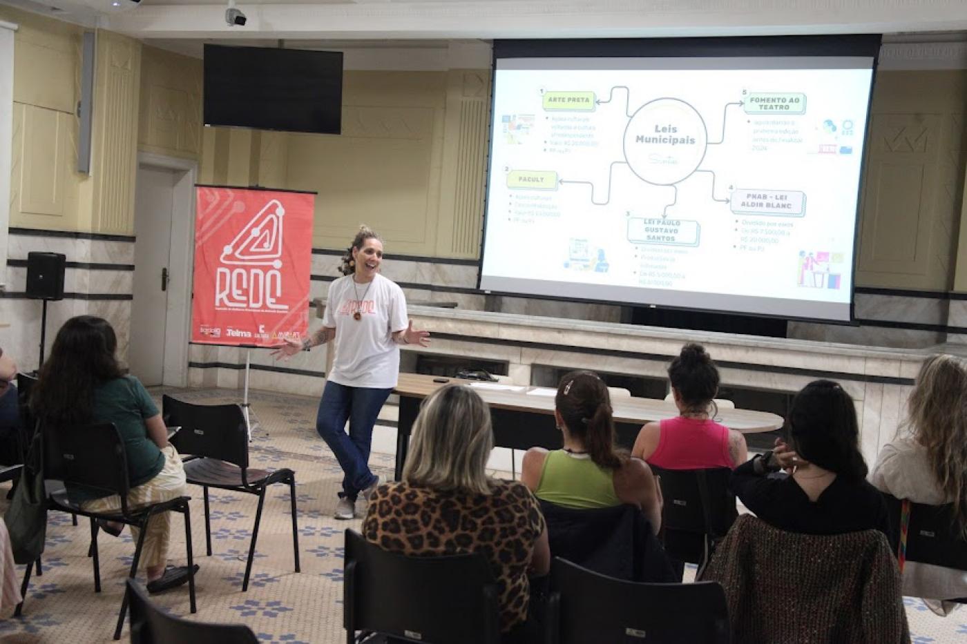 auditório com mulheres sentadas assistindo a uma palestra de uma mulher que está em pé. Há um telão ao fundo. #paratodosverem 