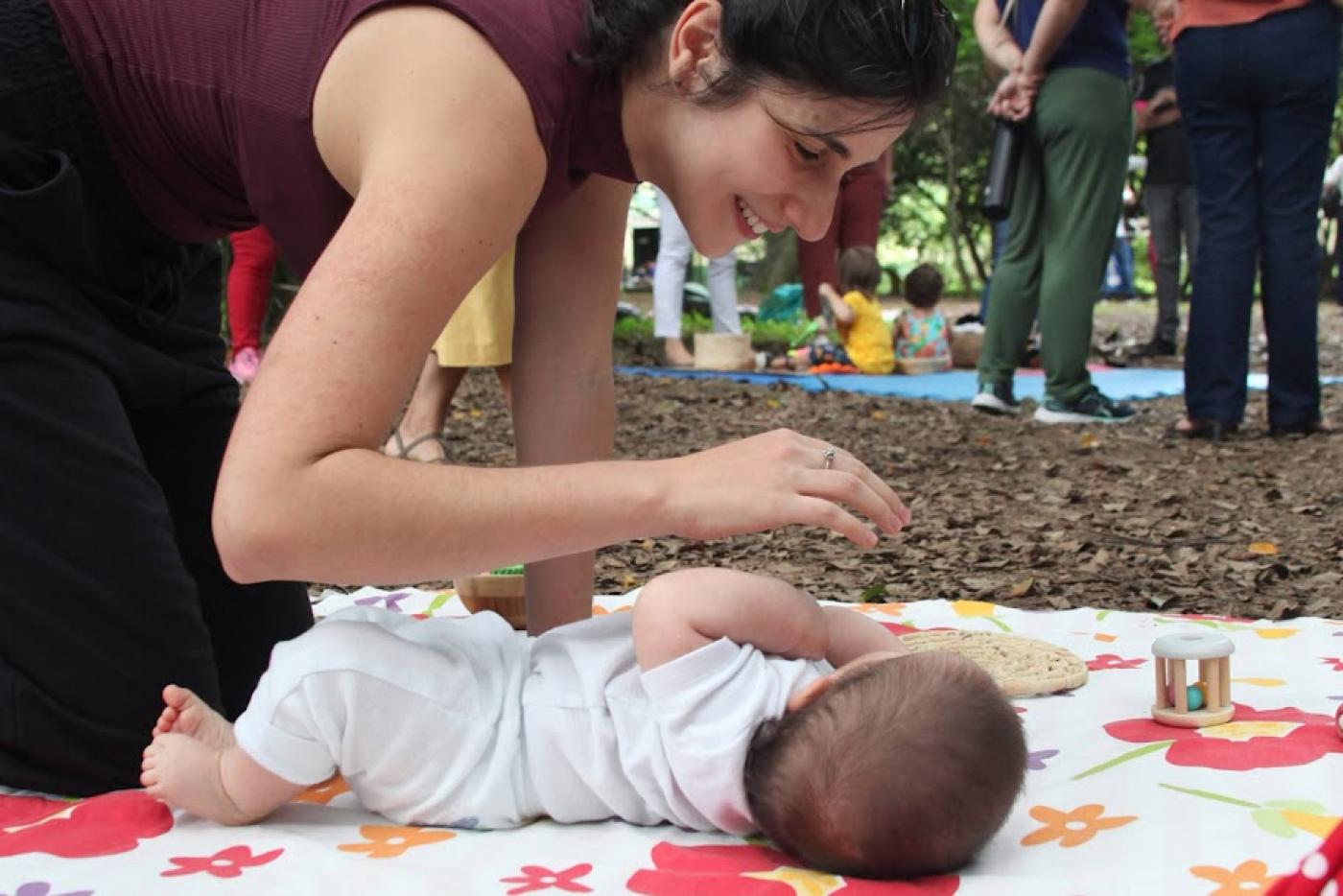 bebê está deitado no chão sobre colchonete. Mãe está ajoelhada olhando para ele e dirigindo uma das mãos ao rosto da criança. #paratodosverem 