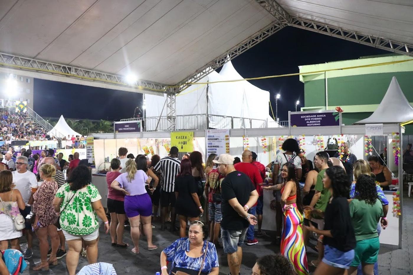 área de alimentacão com pessoas sentadas e pé próximas a mesas e barracas de lanches. #paratodosverem 