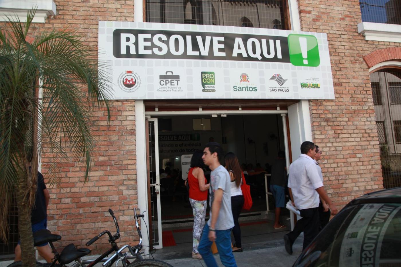 Pessoas passam pela calçada em frente à fachada do Resolve Aqui, indicado em letreiro. #pracegover