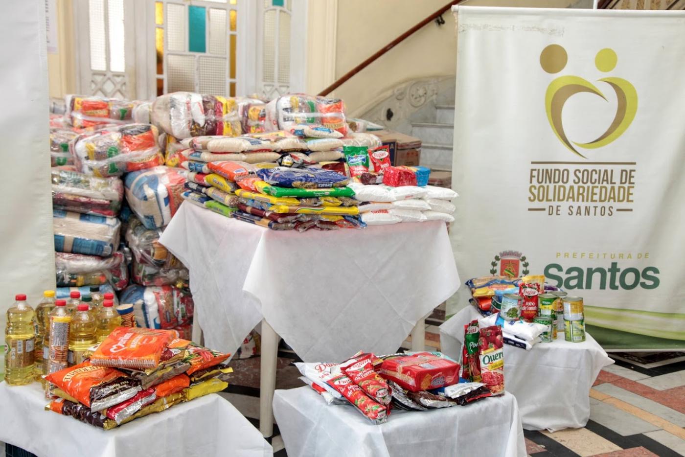 vários alimentos empilhados e dispostos sobre mesas. Ao fundo, um banner do Fundo Social de Solidariedade. #paratodosverem