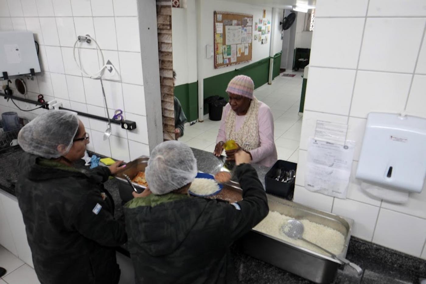 mulheres atrás de balcão servem comida no prato a uma outra mulher. #paratodosverem