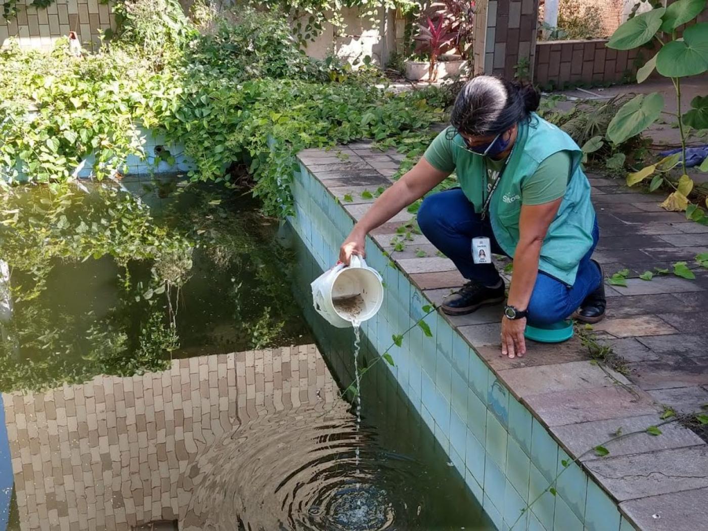 Mulher está agachada à beira de piscina. Ela segura na mão direita uma jarra com um liquido e pequenos peixo e os verte para a piscina. #Paratodosverem