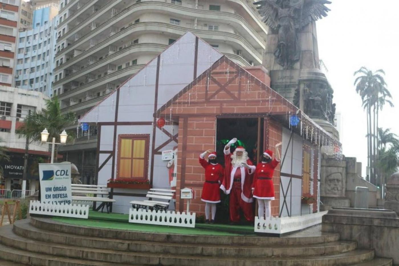 papai noel e mamães noel na frente da casinha na praça #paratodosverem