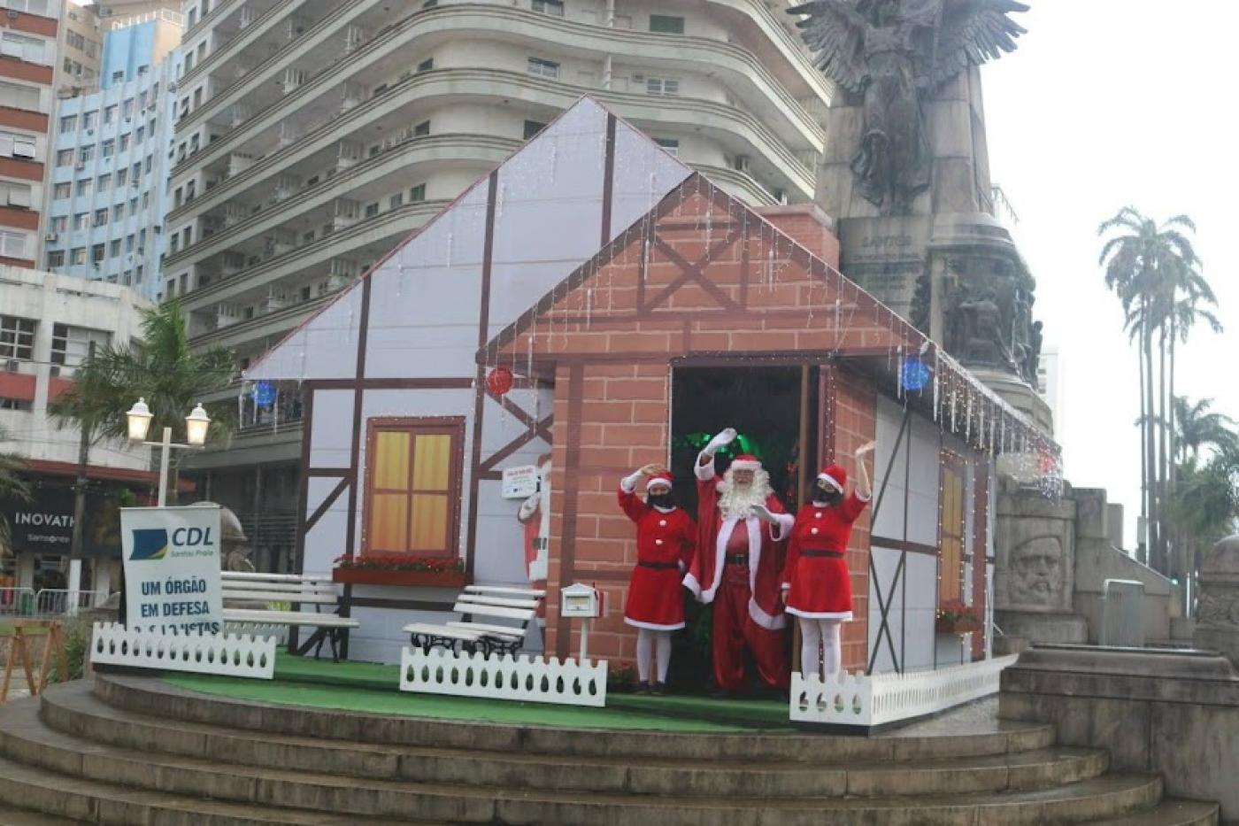 Papai e mamãe noel estão acenando na frente da casinha, no meio de uma praça. #psaratodosverem