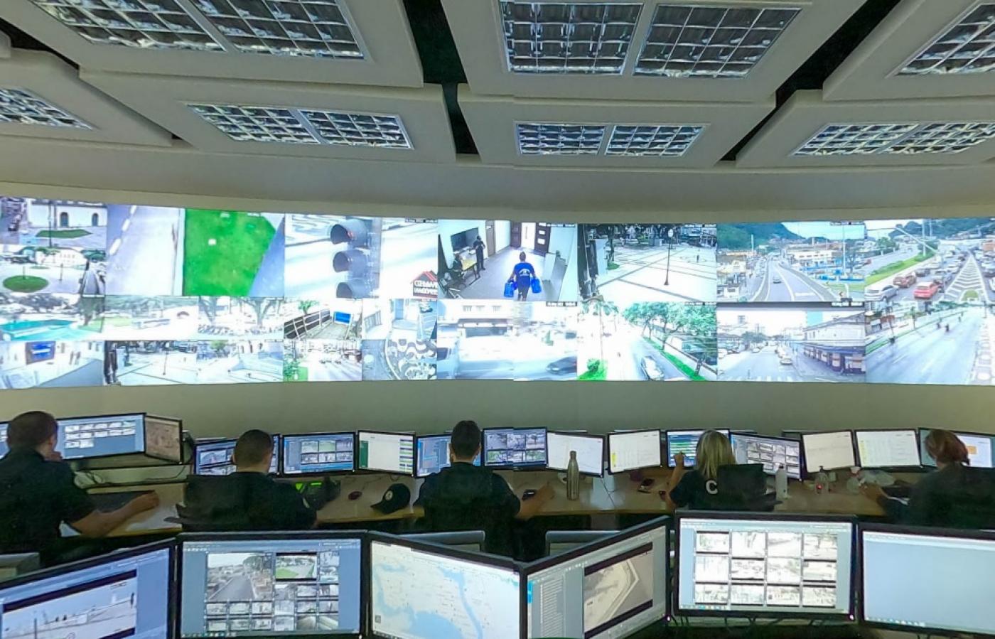 sala do CCO com telas de computadores em primeiro plano, todas com pequenas imagens de monitoramento. Ao fundo, agentes estão olhando outras telas de computador. Mais ao fundo, um grande telão também de monitoramento de regiões da cidade. #paratodosverem