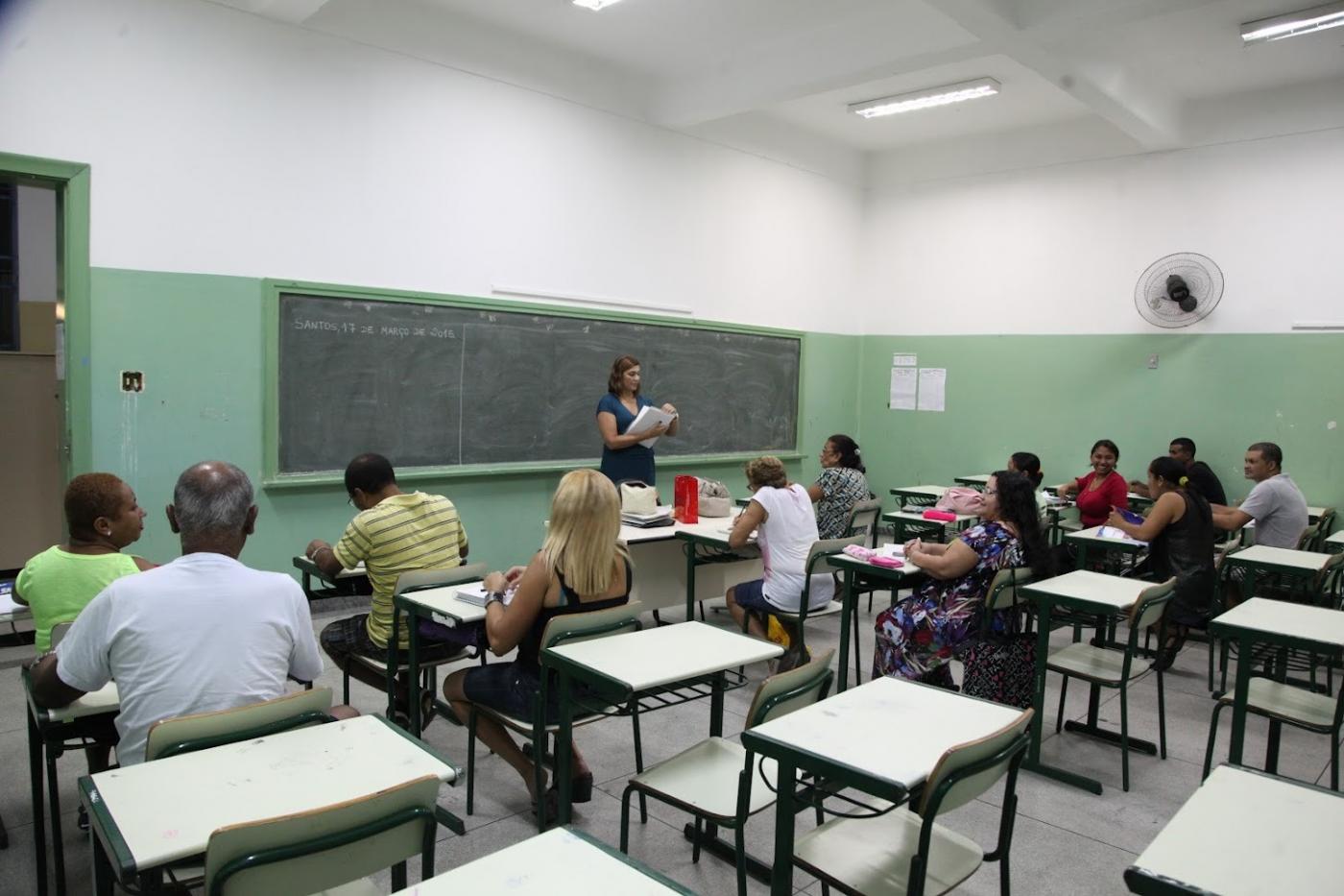 alunos adultos estão sentados em sala de aula. Professora está em pé falando ao grupo. #paratodosverem